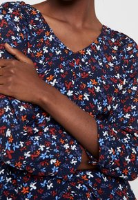 Blommönstrad klänning i marinblått med röda, vita och ljusblå blommor. V-ringning och långa ärmar. Lättviktsmaterial med lätt glans.