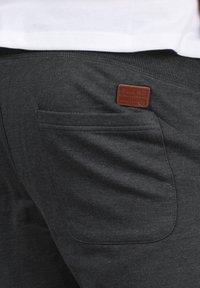 Pantalons de survêtement gris foncé avec une poche à l'arrière, arborant un patch de marque en cuir marron étiqueté "BLEND" près de la taille. Texture douce.