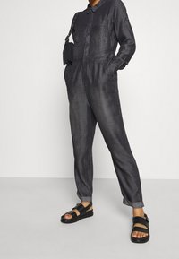 Mono de mezclilla oscura con cuello, mangas largas, dos bolsillos en el pecho y cintura elástica. Combinado con sandalias planas negras.