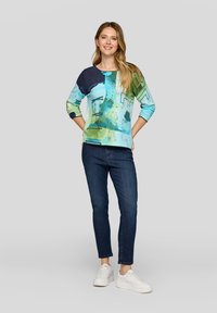 Eine Frau steht lächelnd da und trägt ein langärmliges Oberteil mit abstraktem blau-grünem Muster, dunkellblaue Jeans und weiße Turnschuhe vor einem hellgrauen Hintergrund.