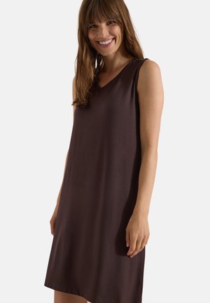 Mujer sonriente con cabello castaño que lleva un vestido sin mangas, marrón oscuro y hasta la rodilla, sobre un fondo liso y claro.
