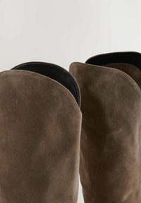 Botas altas de ante marrón con una textura suave, que presentan un borde superior redondeado y forro negro. Sin herrajes ni patrones visibles.