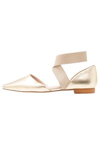 Zapato plano metálico dorado con puntera afilada y una tira elástica beige en el tobillo. Presenta un bajo tacón de madera sólida y una textura lisa.