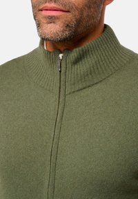 Pull en maille vert olive avec un col côtelé haut et une fermeture éclair argentée au niveau du cou. Texture douce avec une finition lisse.