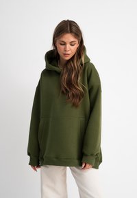 Sweat à capuche vert olive avec une poche avant, coupe oversize et cordon de serrage. Présente des poignets et un ourlet côtelés pour une finition ajustée.