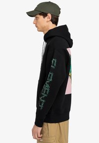 Sudadera negra con bolsillo tipo canguro, con texto verde a lo largo de la manga y un gráfico colorido en la parte posterior sobre un fondo rosa.