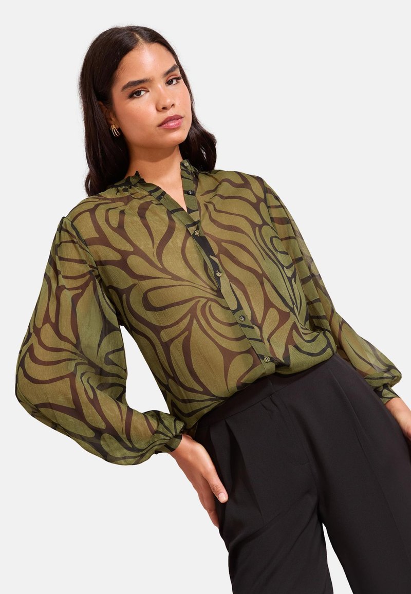 Mlada ženska, oblečena v prosojno olivno zeleno cvetlično bluzo z napihnjenimi rokavi in črne hlače z visokim pasom, stoji s postavljenimi rokami na bokih pred belim ozadjem.