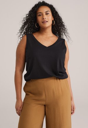 Femme aux cheveux bouclés portant des boucles d'oreilles créoles dorées, un haut noir sans manches à col en V, et un pantalon marron taille haute sur un fond uni.