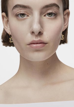 Pendientes de oro con un diseño en forma de oso, que presentan una textura suave y un contorno en contraste. El modelo tiene el cabello corto y un tono de piel neutral.