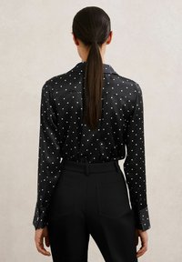 Mustast satiinist särk valgete täppidega, pikkade varrukatega, nööbitavate mansettidega ja tagumise yoke-disainiga, kombineeritud kõrge vöökohaga mustade pükstega.