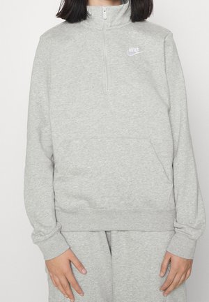 Persoon draagt lichtgrijze Nike kwart-ritssweater en bijpassende joggingbroek, staat met ontspannen handen langs het lichaam.
