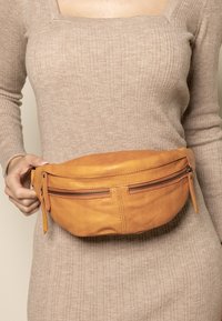 Žena u bež haljini s rebrastim uzorkom drži smešnu kožnu torbicu oko struka s dva zatvarača na razini struka.