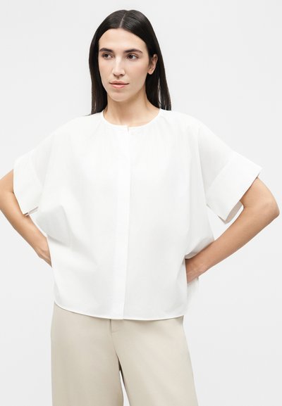 Γυναίκα με μακριά σκούρα μαλλιά, φορώντας άσπρο φαρδύ κοντομάνικο μπλουζάκι και μπεζ παντελόνι, στέκεται με τα χέρια στη μέση, κοιτάζοντας προς τα αριστερά.