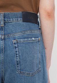 Jean en denim bleu taille haute avec une finition texturée, présentant un patch en cuir foncé à la taille et un design classique à cinq poches.