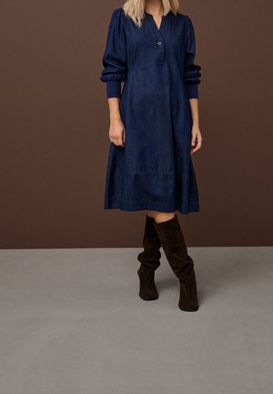Femme portant une robe midi en jean bleu foncé à manches longues bouffantes et des bottes noires molles montantes jusqu'aux genoux.
