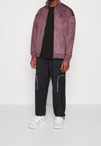 Lila mockaskinnsjacka med dragkedja och svarta ränder, kombinerad med svarta cargo-byxor med gråa detaljer och vita sneakers med texturerade detaljer.