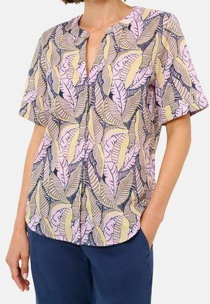 Kvinde iført en kortærmet bluse med mønster af blade i pink, gul og beige, kombineret med mørkeblå bukser.