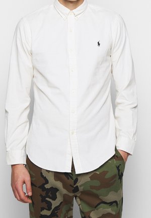 Mann trägt ein weißes, geknöpftes Hemd mit kleinem Logo und grün-braune Camouflage-Hose, Hand in der Tasche, vor schlichtem Hintergrund.