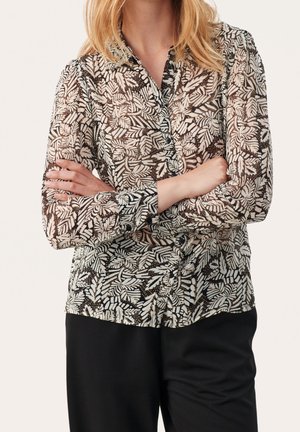 Personne aux bras croisés portant une chemise boutonnée à manches longues avec un motif de feuilles noir et blanc et un pantalon noir.