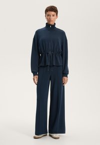 Mornarsko lahek trening komplet, ki vključuje jakno z zadrgo in elastiko v pasu ter široke hlače, v kombinaciji s bež športnimi copati.