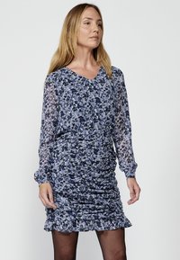 Kukkakuosiset mekko sinivioletin sävyissä, joissa on pitkät hihat, V-kaula-aukko, rypytetty yksityiskohta ja rypytetty helma.