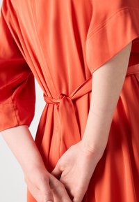 Rochie din satin portocalie cu talie legată, având mâneci largi și o textură netedă. Țesătura se așază ușor, evidențiind un design fluid.