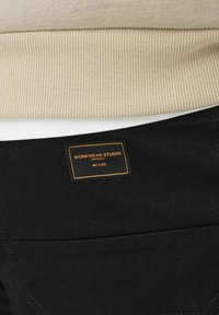 Czarna tkanina o teksturowej powierzchni, z widocznym beżowym ściągaczem i metką z napisem "WORKWEAR STUDIO" w pomarańczowym szwie.