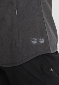 Chemise gris foncé avec des icônes de tissu à séchage rapide et extensible imprimées près de l'ourlet, portée sur un pantalon noir avec une poche zippée.