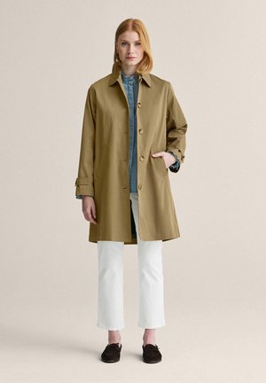 Kvinde med rødt hår iført en tan knælængde frakke over en blå flæset skjorte, hvide bukser og sorte mokkasinsko, stående mod en enkel baggrund.