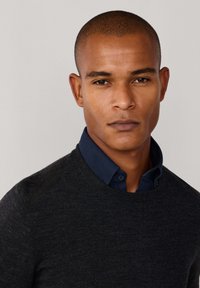 Tmavě šedý svetr s kulatým výstřihem, který je navrstven na námořnické košili s knoflíky. Tkanina vypadá pleteně s hladkou texturou a minimálním zdobením.