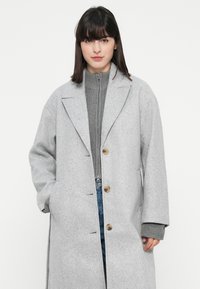 Donna che indossa un lungo cappotto grigio chiaro con bottoni sopra un maglione grigio con zip e jeans blu, in piedi di fronte a uno sfondo bianco semplice.