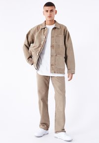 Giacca di denim beige con tasche frontali, T-shirt bianca sotto, e jeans beige abbinati. Il modello indossa sneakers bianche. Stile semplice e casual.