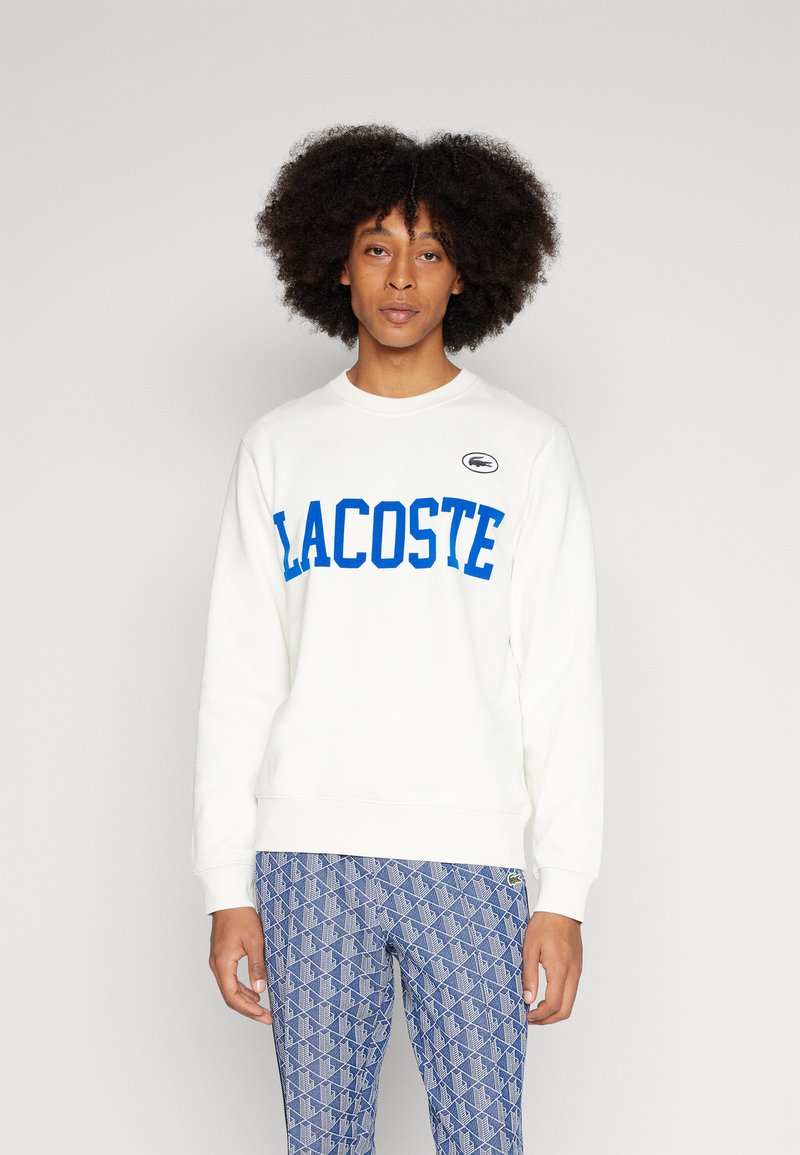 Felpa bianca con lettering azzurro "LACOSTE" e piccolo logo ricamato, colletto a giro, polsini a costine, abbinata a pantaloni blu con motivo.
