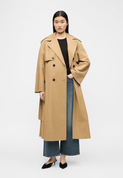 Γυναίκα που στέκεται με το χέρι στην τσέπη, φορώντας μακρύ μπεζ παλτό τύπου trench, μαύρο τοπ, φαρδιά μπλε τζιν και μαύρα παπούτσια με χαμηλό τακούνι, σε απλό φόντο.