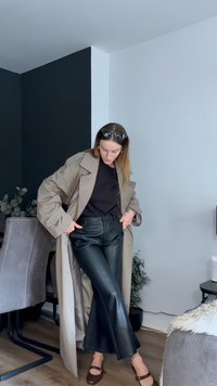 Casaco trench bege sobre t-shirt preta e calças de couro pretas largas. Sapatos de salto castanhos. Modelo em pé, com as mãos nos bolsos, usando óculos de sol.