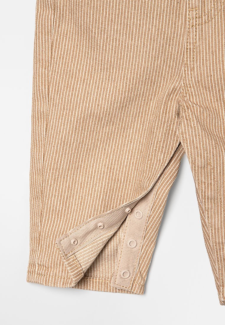 Beige gestreifte Latzhose mit Klappe und Druckknopfverschluss an der Beinöffnung. Hergestellt aus einem strukturierten Stoff. Mit vertikalen weißen Streifen.