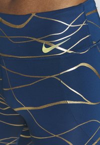Granatowe legginsy o gładkiej fakturze, z metalicznymi złotymi falistymi liniami i małym żółtym logo Nike na talii.