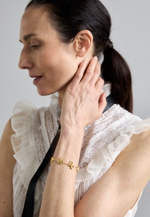 Gullarmbånd med blomstermotiver, båret på håndleddet mot en bakgrunn av en rynket, hvit bluse i blonder med svart bånddetalj.