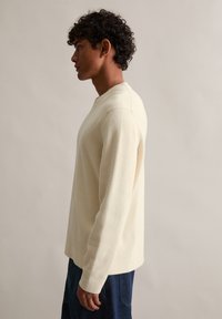 Sweter z długim rękawem i okrągłym dekoltem w fakturowanym kremowym materiale. Luźny krój z minimalną ilością szwów. Połączony z ciemnoniebieskimi dżinsami dla kontrastu.