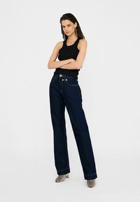 Jean droit en denim bleu foncé avec des coutures contrastées, doté d'une fermeture à boutons et de poches avant. Associé à un débardeur noir.