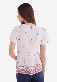 Witte blouse met rode geometrische borduurdetails, korte puffmouwen en een geplooide peplum taille. Zachte, lichtgewicht stof met een gestructureerde afwerking.