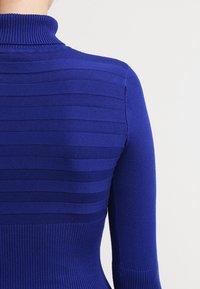 Blauer Strickpullover mit hohem Kragen, der horizontale Streifenmuster und gerippte Textur aufweist. Figurbetonte Silhouette, die sich an der Taille verjüngt.