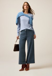 Helles blaues Sweatshirt über die Schultern drapiert, kombiniert mit türkisfarbenen weiten Cordhosen und braunen Knöchelstiefeln. Hält eine braune Handtasche.
