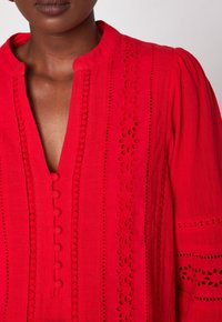 Blusa rossa con scollo a V, caratterizzata da fantasie traforate decorative e dettagli con bottoni. Tessuto morbido con dettagli testurizzati e maniche a sbuffo.