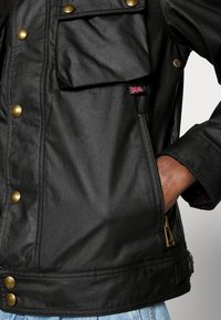 Černá bunda z hladkého materiálu se zlatými patentkami, přední kapsou a zipovým detailem; součástí je malá visačka s motivem vlajky Spojeného království.