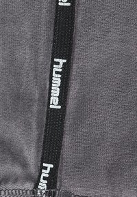 Grauer Stoff mit einer strukturierten Oberfläche, der ein schwarzes Band mit weißem "hummel"-Logo aufweist. Die Nähte sind entlang des Randes sichtbar.