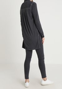 Persona che indossa una lunga maglietta grigio scuro, pantaloni grigio scuro aderenti e sneakers bianche, in piedi contro uno sfondo chiaro e semplice.