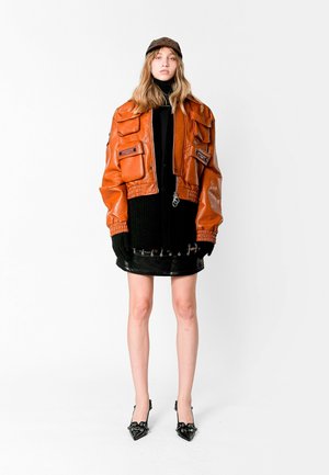 Chaqueta de cuero naranja corta con múltiples bolsillos, un cuello de tortuga negro de punto, una falda corta brillante negra y tacones de punta negra.