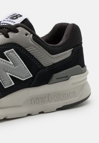 Melnas un pelēkas sporta apavi ar teksturētu augšdaļu, mīkstu apkakli un izteiktu sānu "N" zīmi. Mīksta gumijas zole ar zīmola nosaukumu.