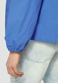 Blau leichte Jacke mit elastischen Bündchen, die eine glatte Textur aufweist. Kombiniert mit hellblauer Jeans, sichtbare Taschenrücken-Details.